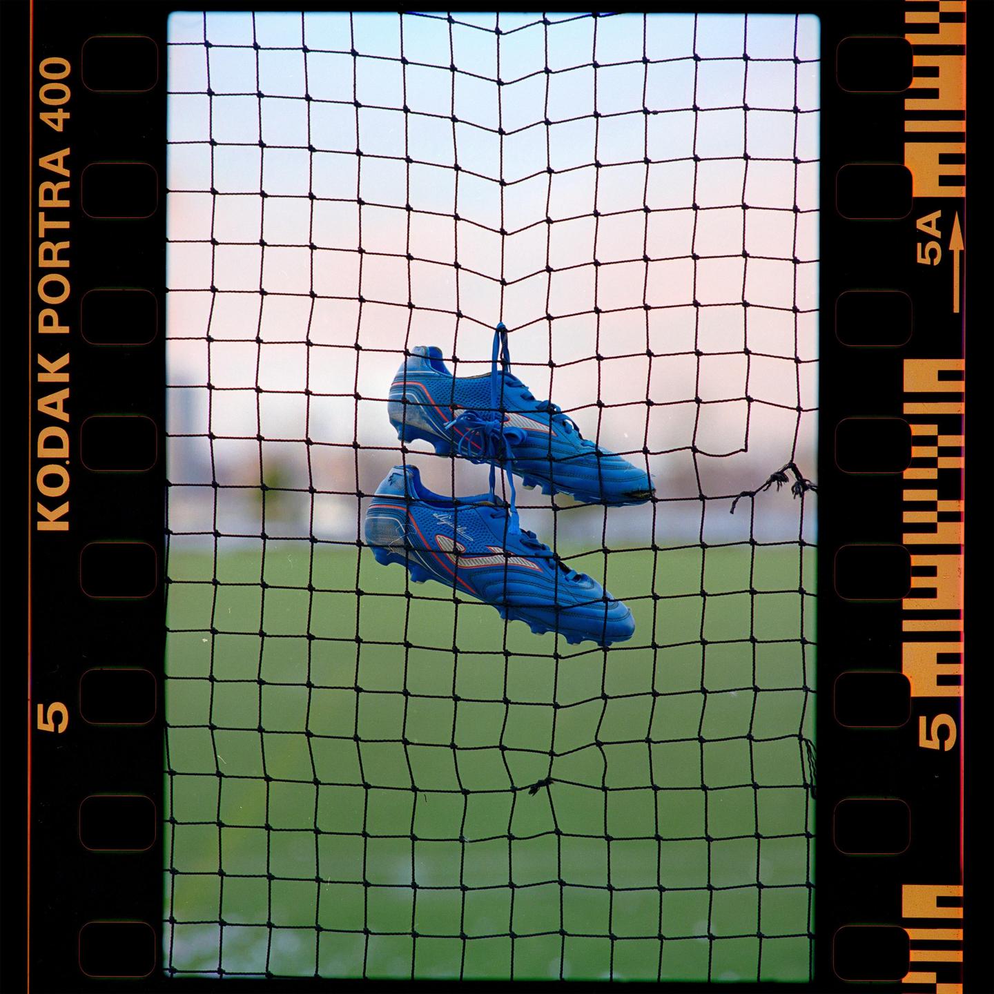 Two blue soccer cleats hang from a net with a green field and sky in the background.
