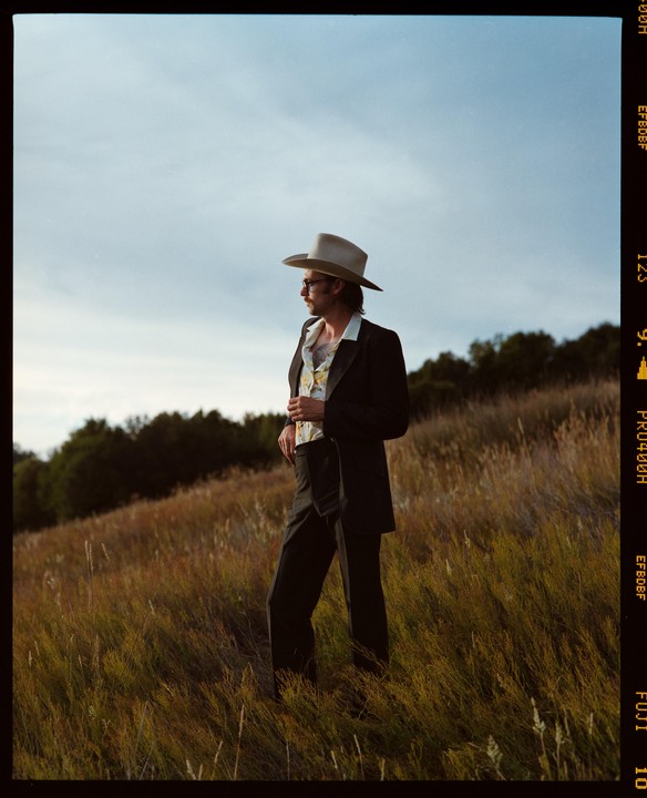 A man in a suit, cowboy hat, and sunglasses stands in a field of tall grass looking to the right.
