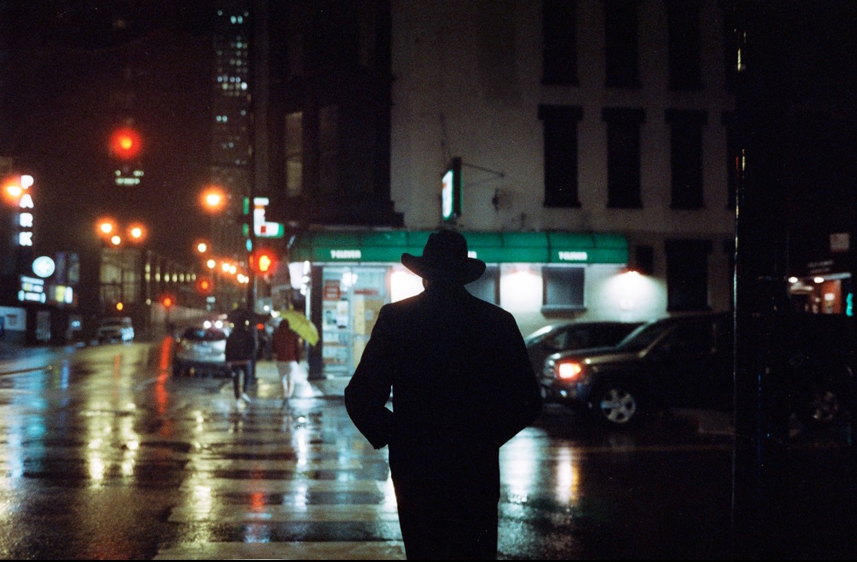A person in a hat and coat stands on a wet city street at night, with reflections of streetlights and cars on the pavement.