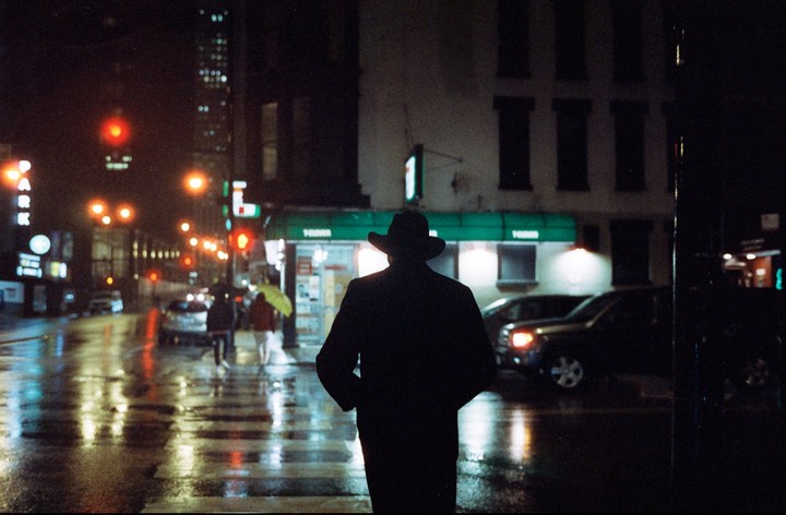 A person in a hat and coat stands on a wet city street at night, with reflections of streetlights and cars on the pavement.