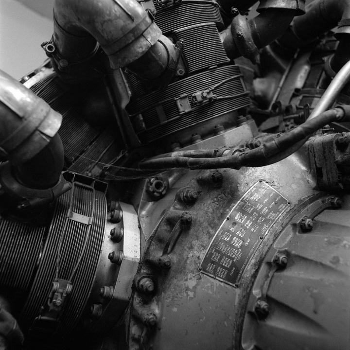 A close-up, black and white photograph shows the intricate details of a large, industrial engine with various pipes, bolts, and a data plate.