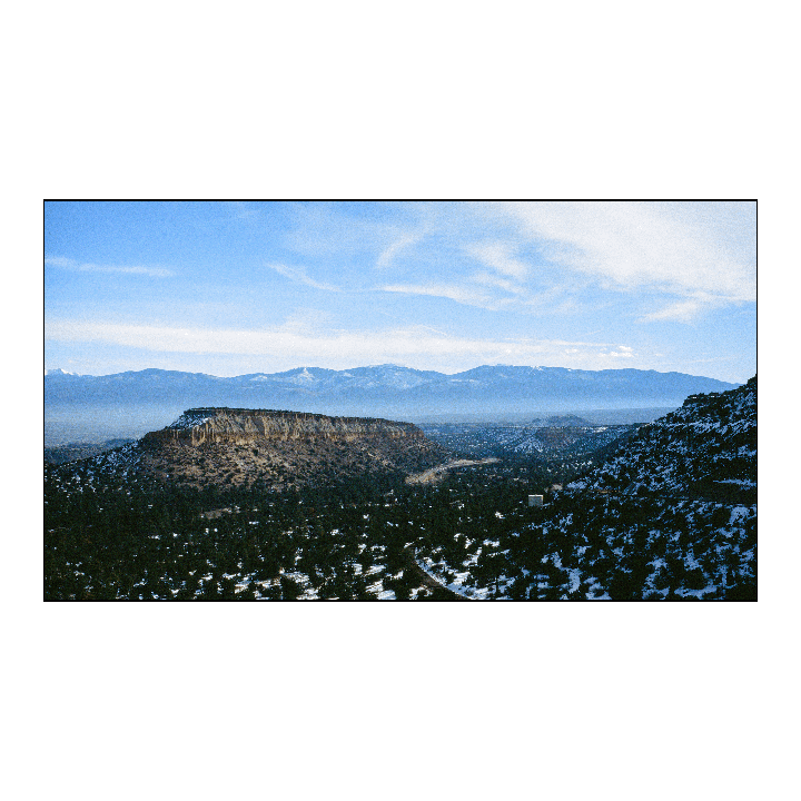A wide landscape shows a valley with a mesa, evergreen trees, and snow-capped mountains under a blue sky.