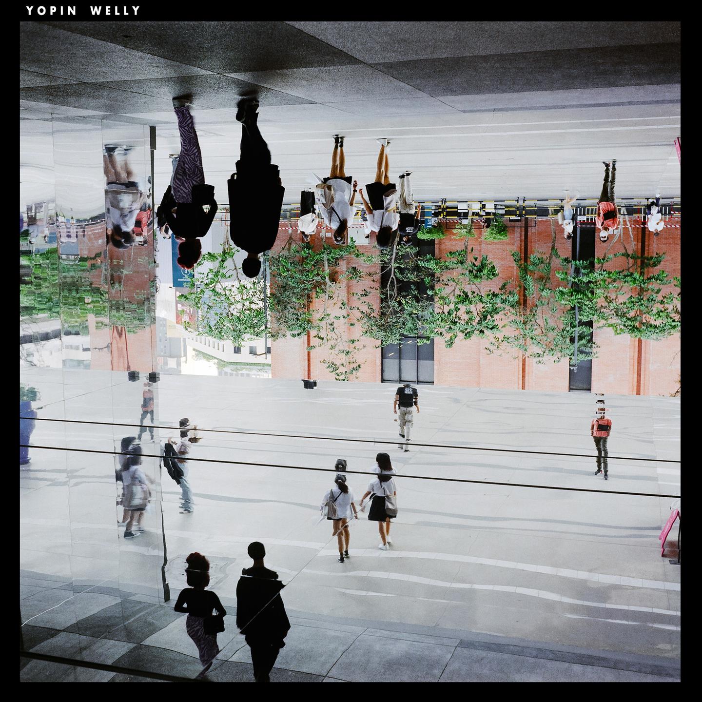 People walk on a street with reflections of buildings and trees on a glass surface.