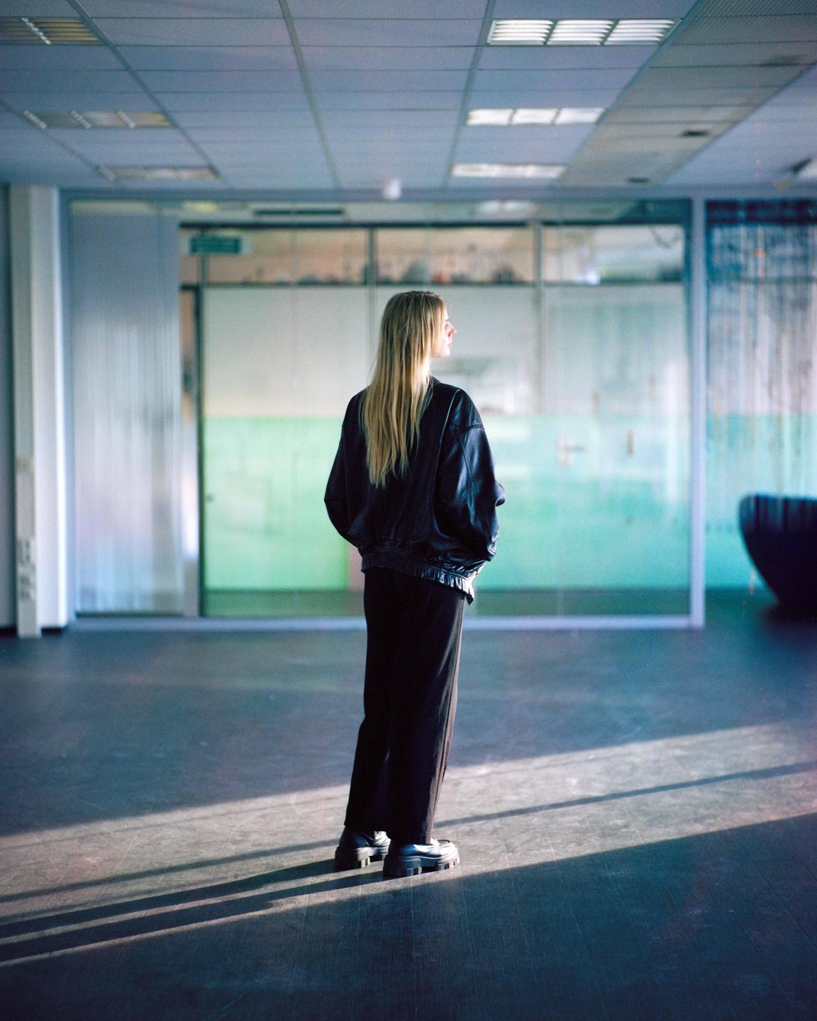 A woman with long blonde hair stands in a room with glass walls and a tiled ceiling, looking to her right.