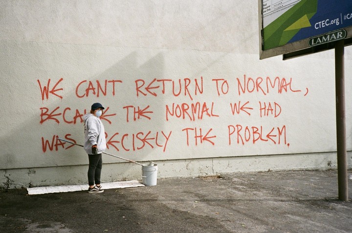 A person wearing a face mask and a baseball cap stands next to a wall with red graffiti that reads "WE CANT RETURN TO NORMAL, BECAUSE THE NORMAL WE HAD WAS PRECISELY THE PROBLEM."