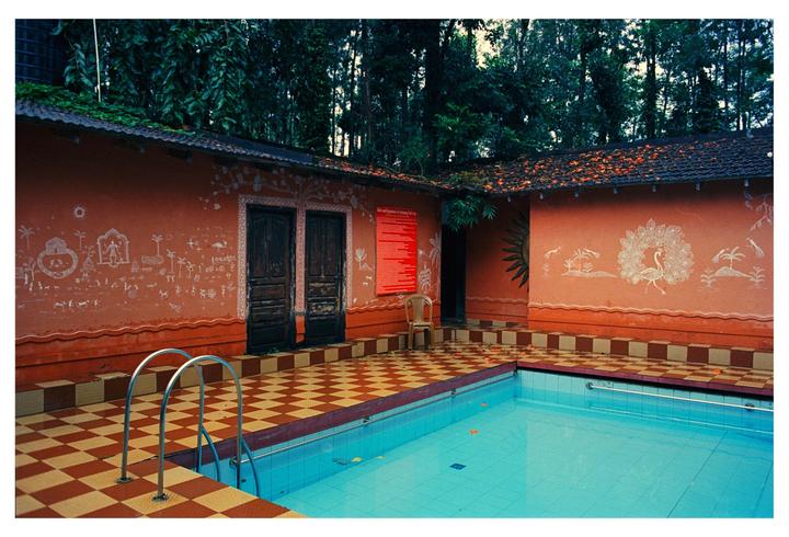 A swimming pool with a checkered tile surround is next to a building with orange walls and white painted designs, with tall trees in the background.