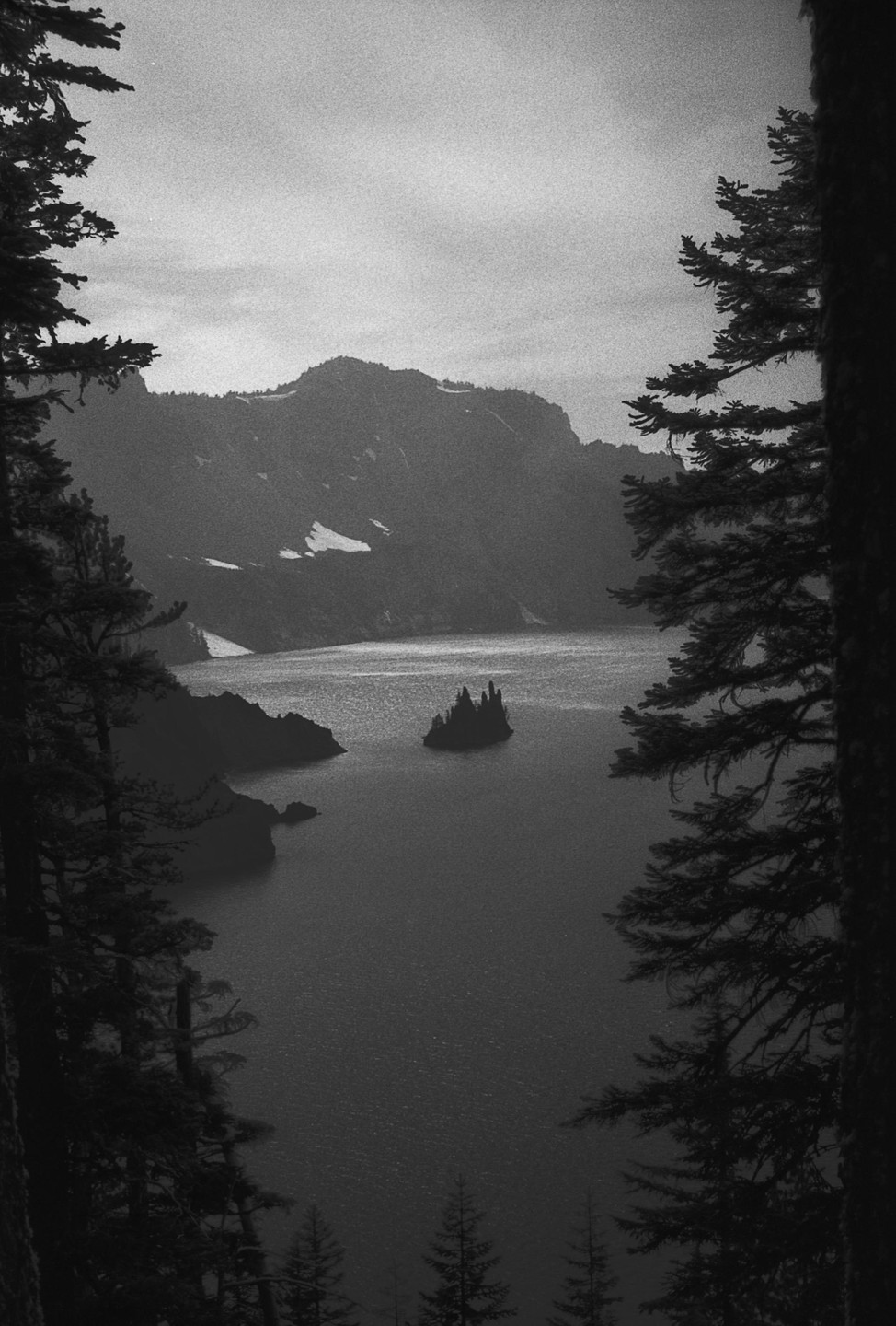 A black and white landscape shows a lake with a small island in the middle, framed by tall pine trees on either side, with snow-capped mountains in the background.