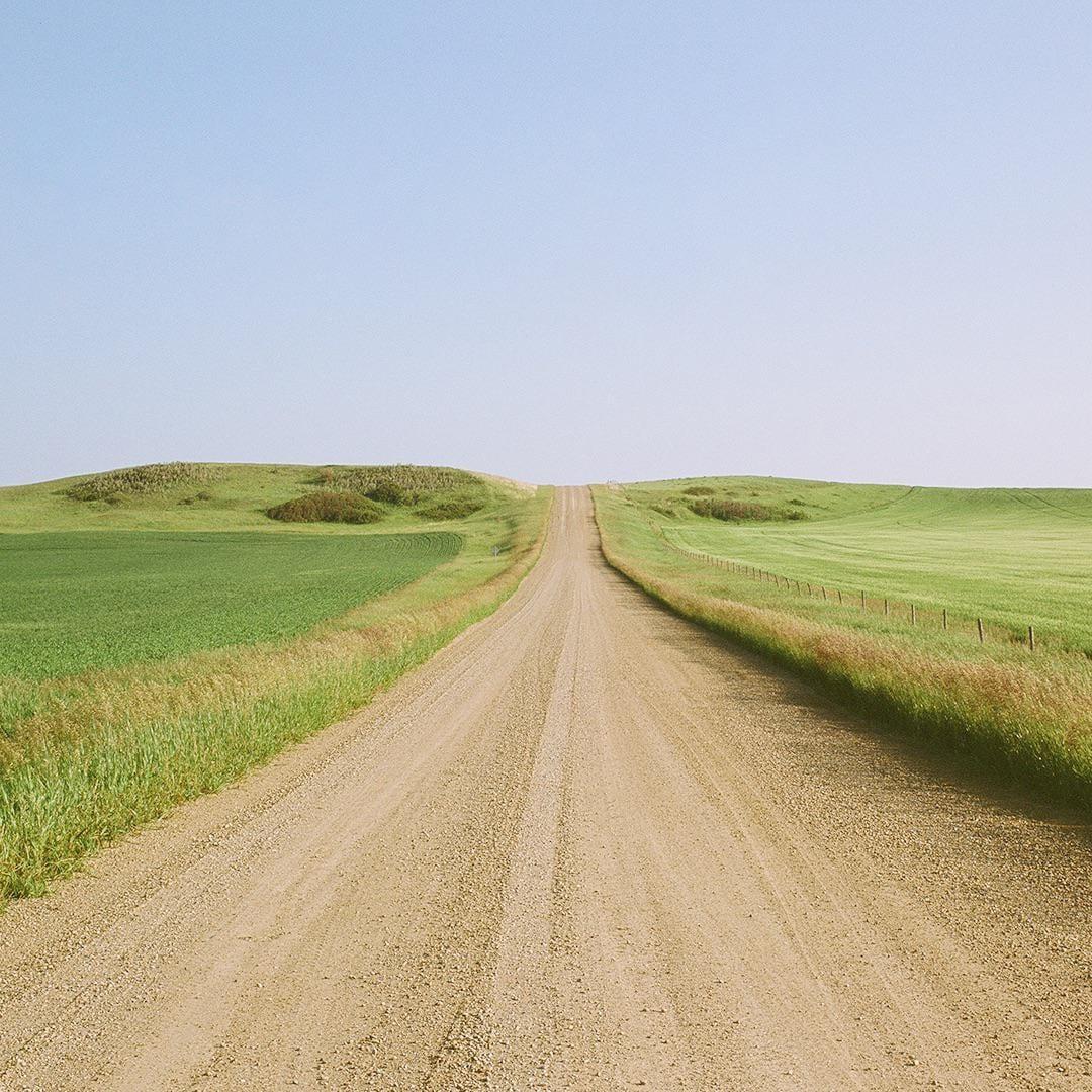 A long gravel road stretches into the distance, flanked by green fields and rolling hills under a clear sky.