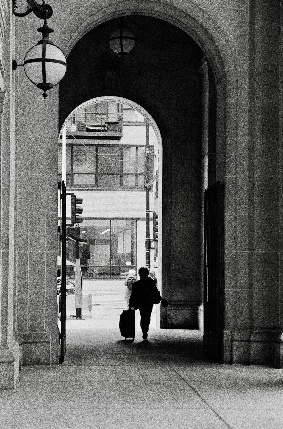 A person pulls a suitcase through an arched walkway, with a street and buildings visible in the background.
