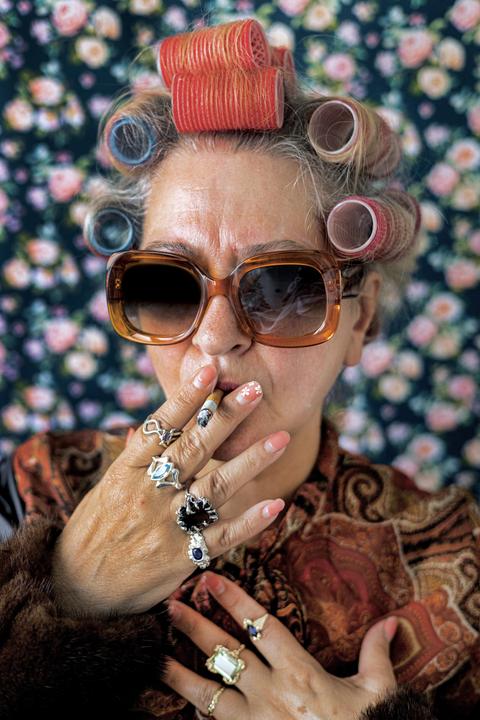 A woman with hair curlers and sunglasses smokes a cigarette against a floral background.