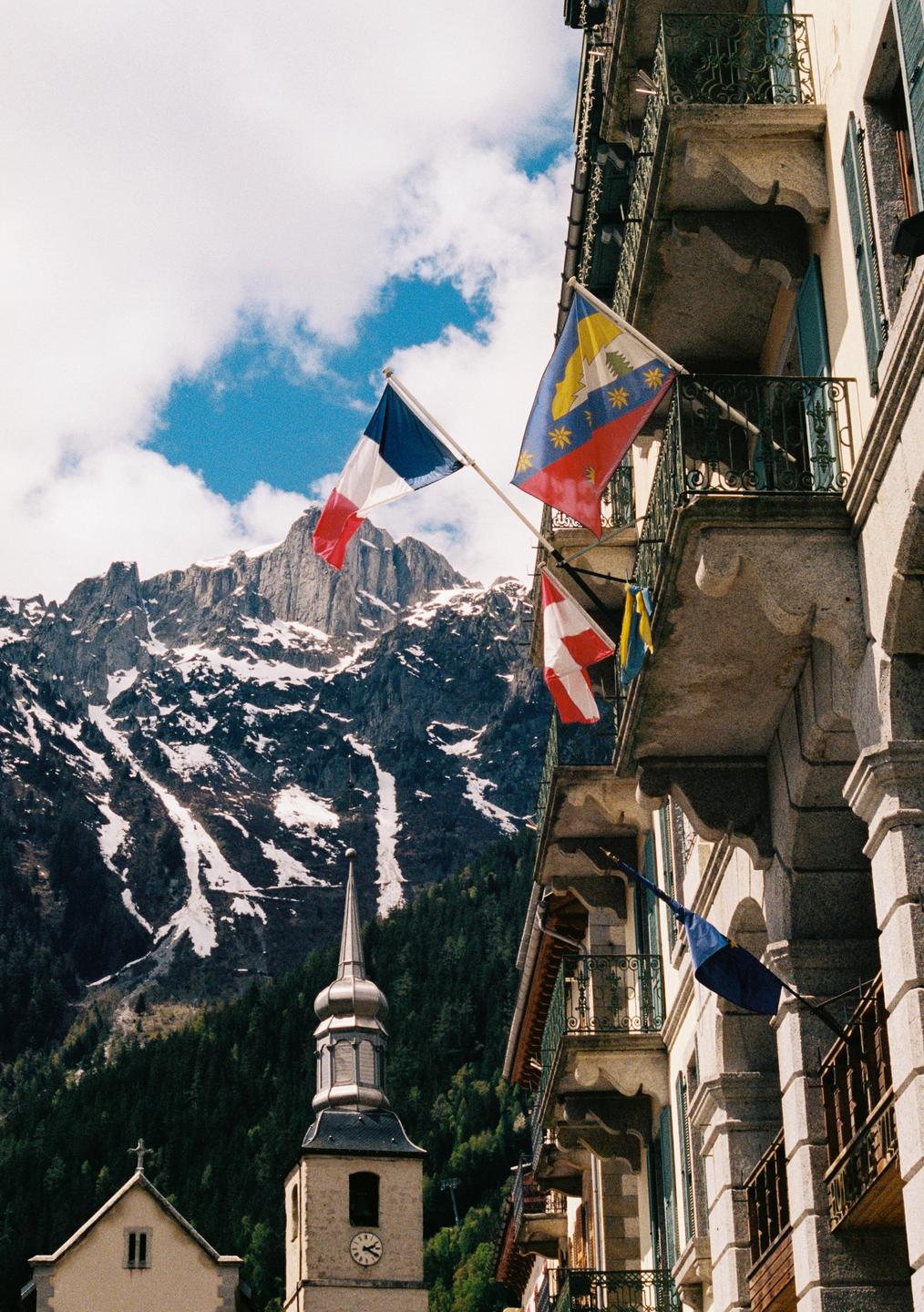 Flags hang from balconies of a building with a church steeple and snow-capped mountains in the background.