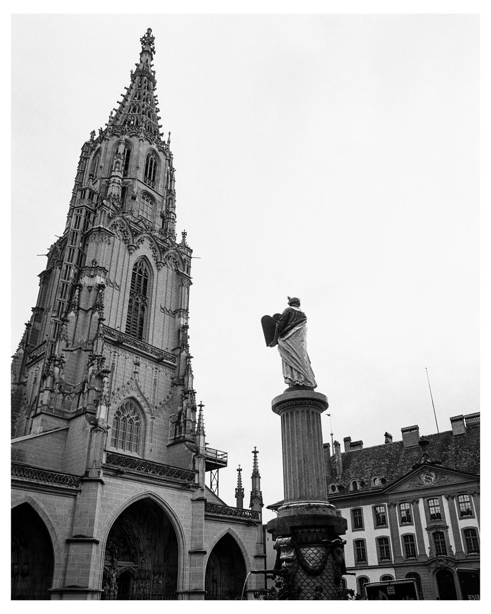 A tall gothic cathedral spire stands next to a statue on a column in a city square.