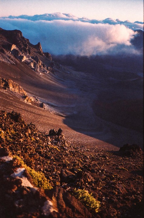A vast volcanic landscape is illuminated by the rising sun, with clouds filling the valley below.