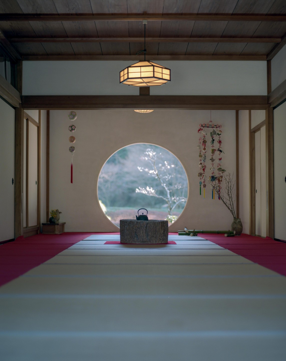 A black teapot sits on a wooden stump in front of a circular window looking out onto a tree.