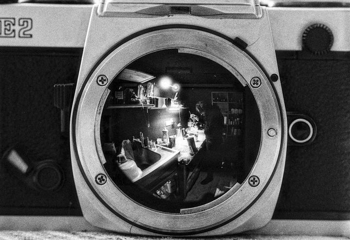 A person works in a darkroom, reflected in the lens mount of a camera.