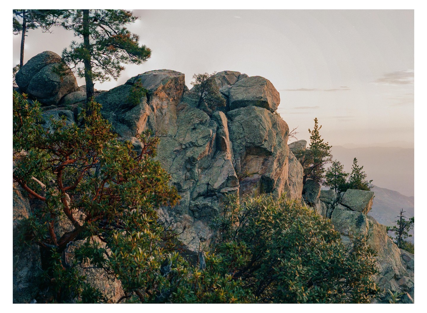 A rocky mountain peak is covered with trees and bushes under a pale sky.