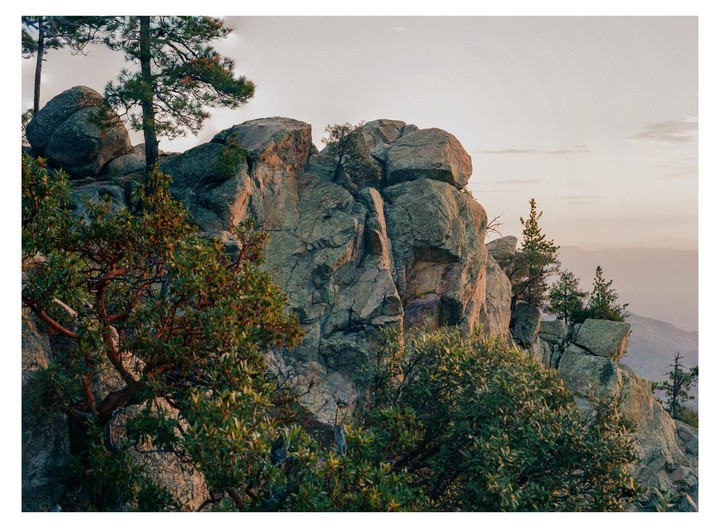 A rocky mountain peak is covered with trees and bushes under a pale sky.