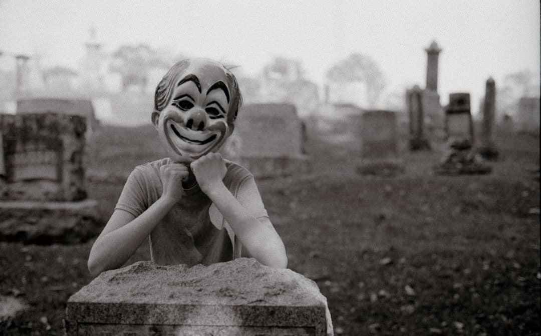 A person wearing a clown mask rests their chin on their fists in a cemetery.