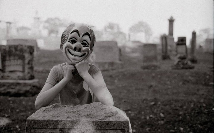 A person wearing a clown mask rests their chin on their fists in a cemetery.