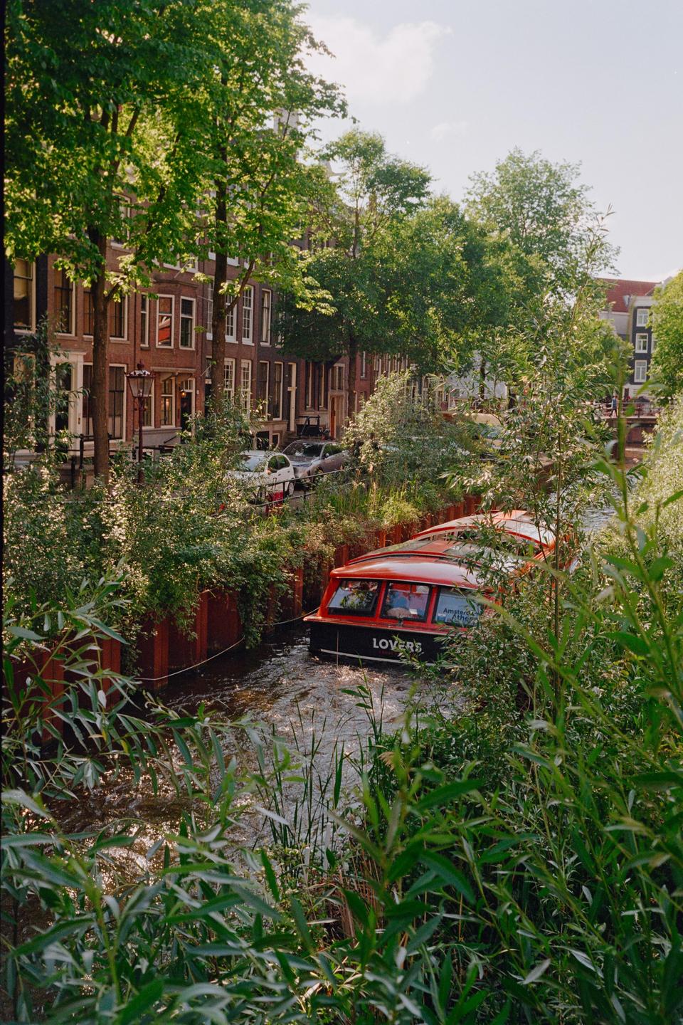 A red and black boat with "LOVERS" written on its side navigates a narrow canal surrounded by lush green foliage and historic buildings.