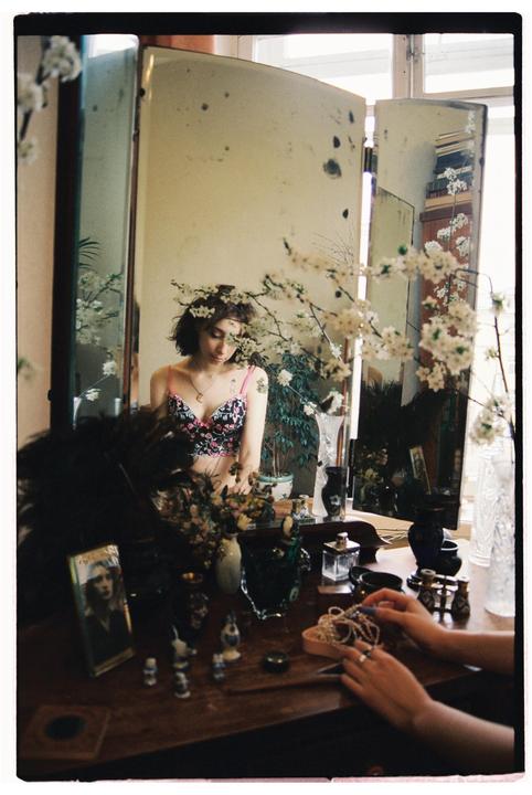 A woman in a floral bra is reflected in a three-panel mirror, surrounded by white blossoms and various trinkets on a wooden dresser.
