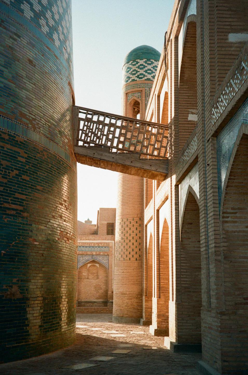 A narrow alleyway is visible between two brick buildings, with a wooden bridge connecting them and a domed tower in the background.