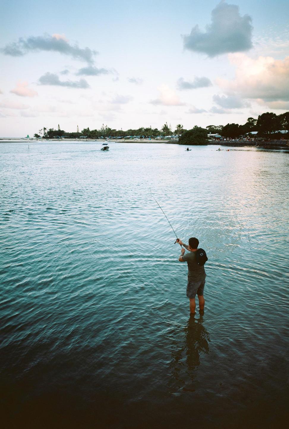 A man stands in the water fishing with a rod and reel, with a shoreline and boats in the background.