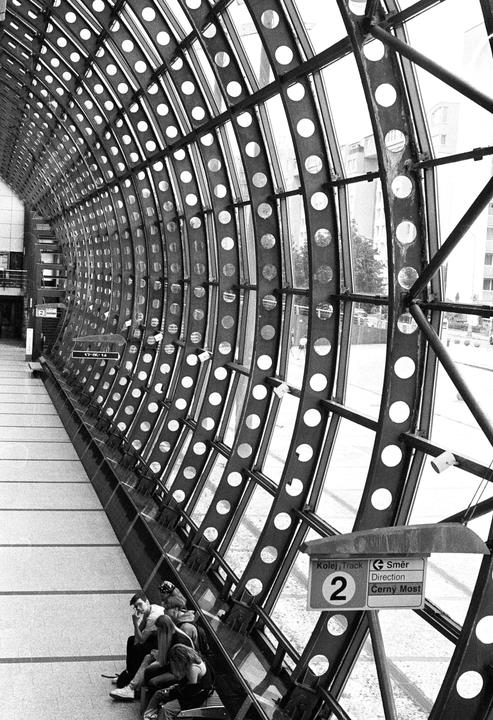 A group of people sit on a bench next to a large glass and metal structure with a sign for "Cerny Most" in the foreground.