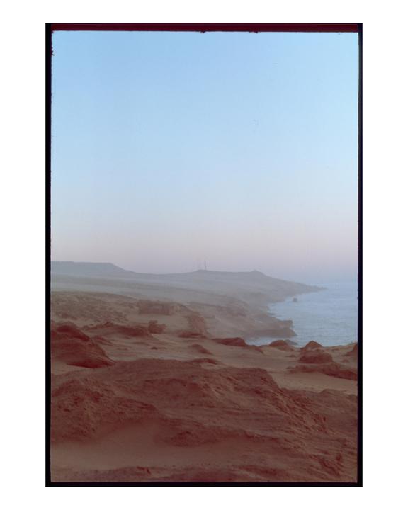 A hazy coastal landscape features a rocky shoreline meeting the ocean under a pale sky.