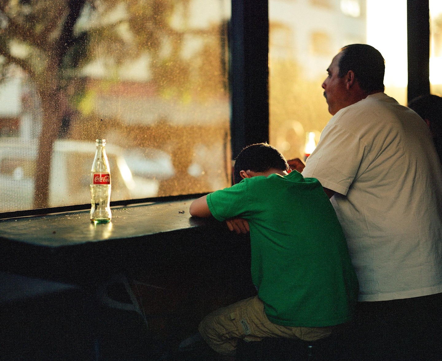 A man and a boy sit at a counter next to a window with a Coca-Cola bottle on the counter.