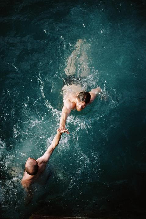 Two men are reaching for each other's hands in dark blue water.