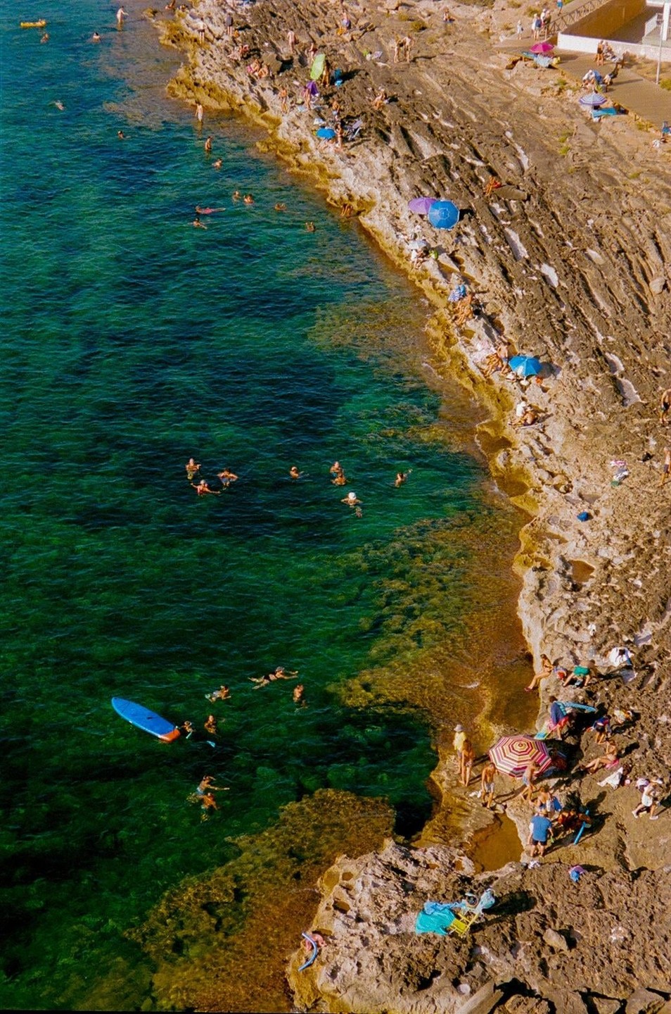 A high-angle view shows people swimming in the clear turquoise water next to a rocky beach where others are sunbathing under umbrellas.