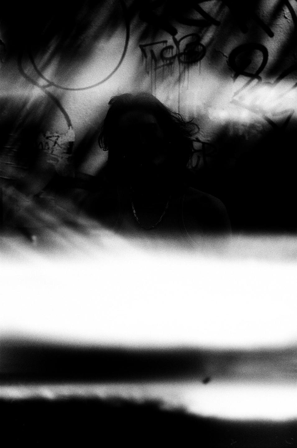 A person with long hair and a necklace is silhouetted against a wall with graffiti.