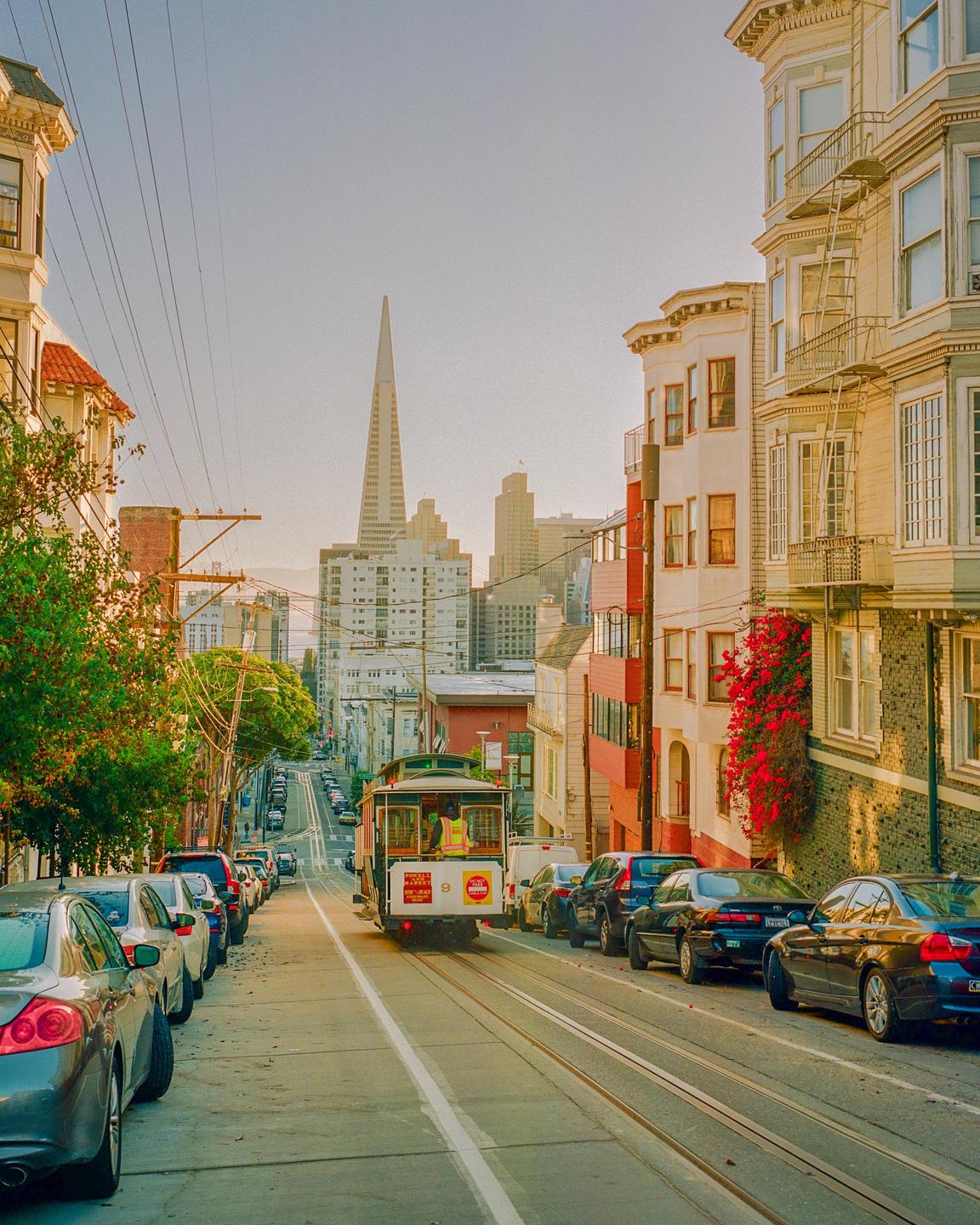 A cable car travels down a steep street lined with parked cars and buildings, with a tall, pointed skyscraper in the distance.