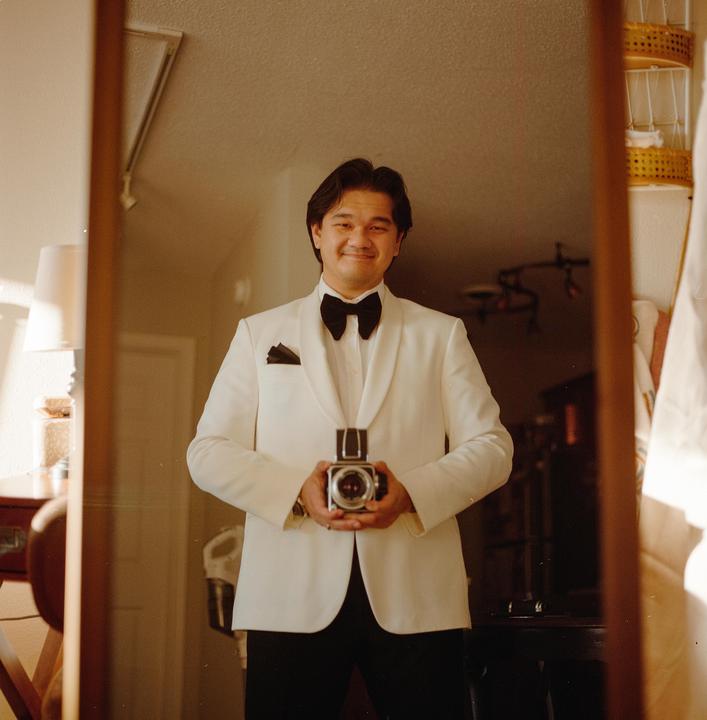 A man in a white tuxedo jacket and black bow tie holds a camera and smiles at his reflection in a mirror.