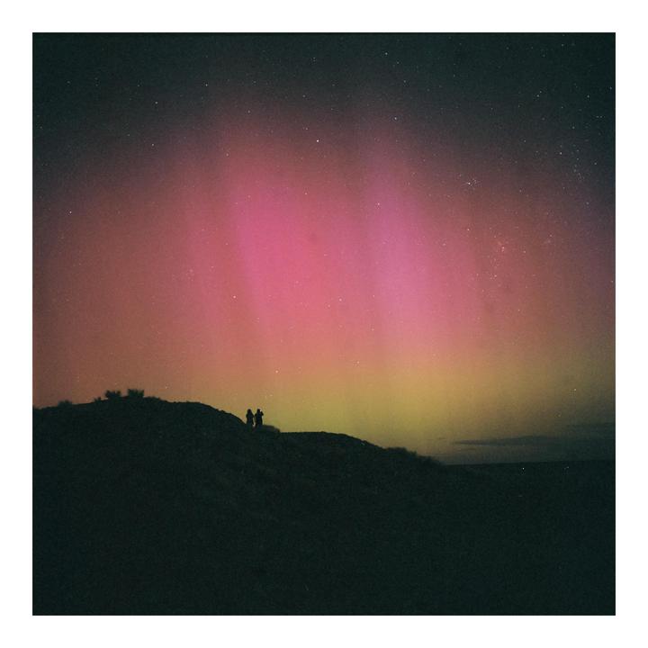 Two people stand on a dark hill silhouetted against the vibrant pink and yellow hues of the aurora australis and a starry night sky.