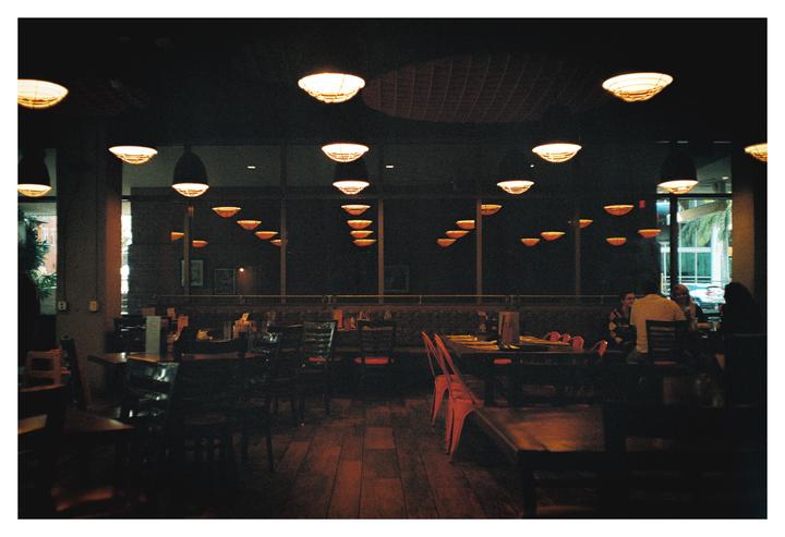 A restaurant interior is dimly lit by numerous ceiling lights and features tables and chairs, with a few patrons visible in the background.