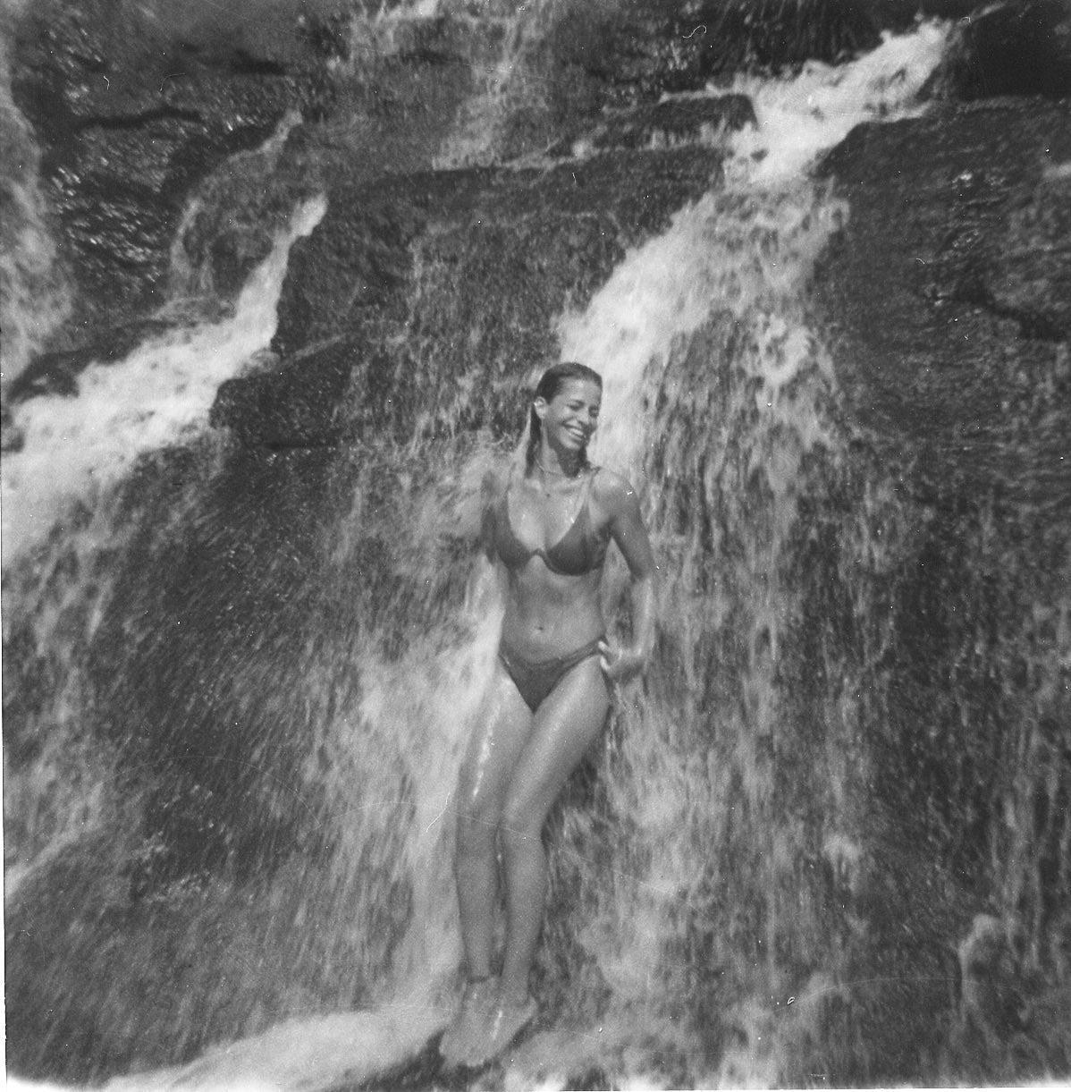 A woman in a bikini stands smiling under a waterfall.