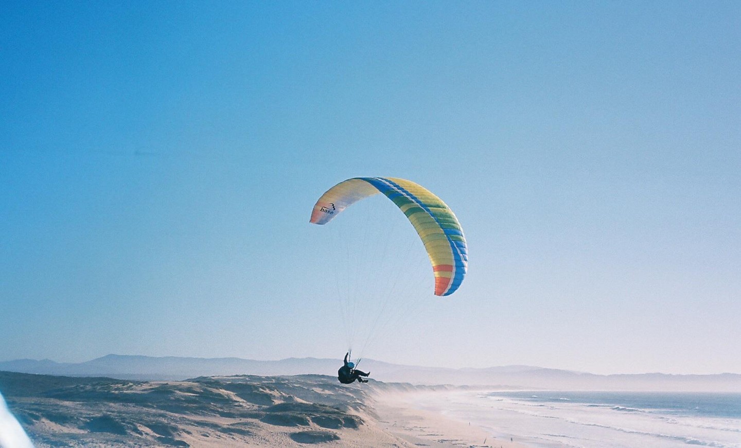 A paraglider soars above a sandy beach with waves crashing on the shore and mountains in the distance.