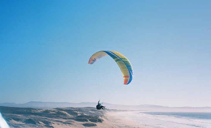 A paraglider soars above a sandy beach with waves crashing on the shore and mountains in the distance.