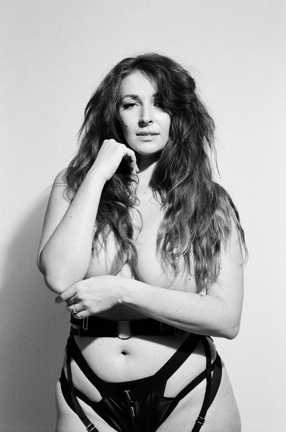 A woman with long, wavy hair poses topless, wearing only a black harness and garter belt.