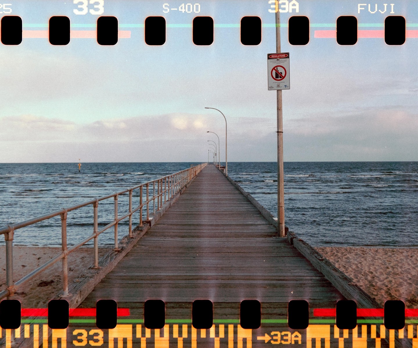 A long wooden pier with metal railings extends into the ocean under a cloudy sky.