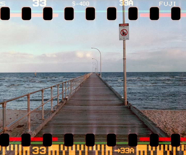 A long wooden pier with metal railings extends into the ocean under a cloudy sky.