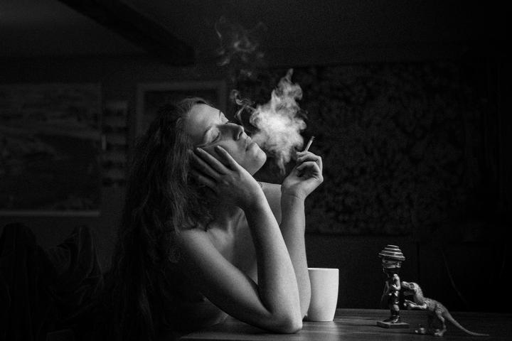 A woman with closed eyes exhales smoke from a cigarette while sitting at a table with a mug and a dinosaur figurine.