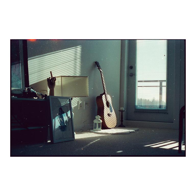 Sunlight streams through blinds onto a guitar, lamp, and framed picture in a room.