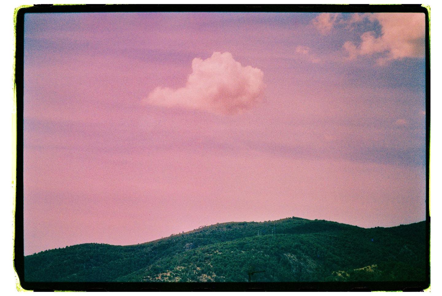 A single cloud floats in a pink sky above a green mountain.
