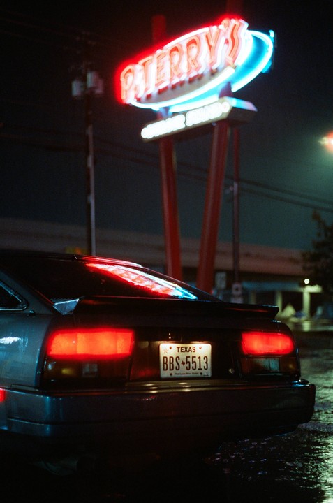 The rear of a car with its brake lights on is parked in front of a neon sign at night.
