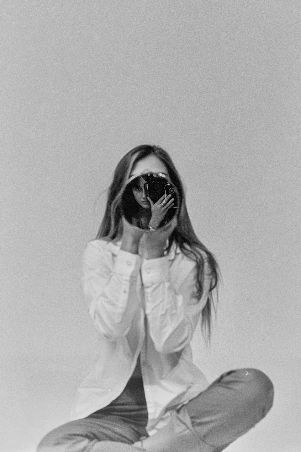 A woman sits holding a round mirror that reflects her face and a camera.