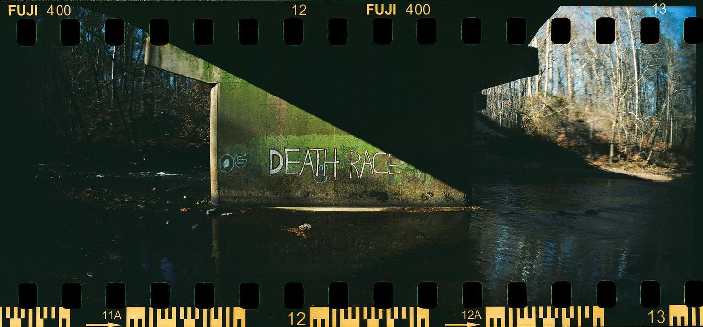 A concrete bridge with graffiti reading "DEATH RACE" casts a shadow over a river, with a forest visible in the background.