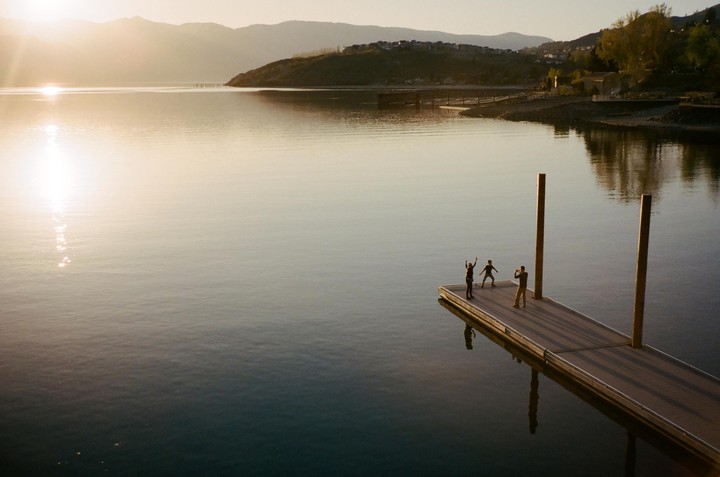 Three people stand on a dock over a lake with mountains in the background at sunset.